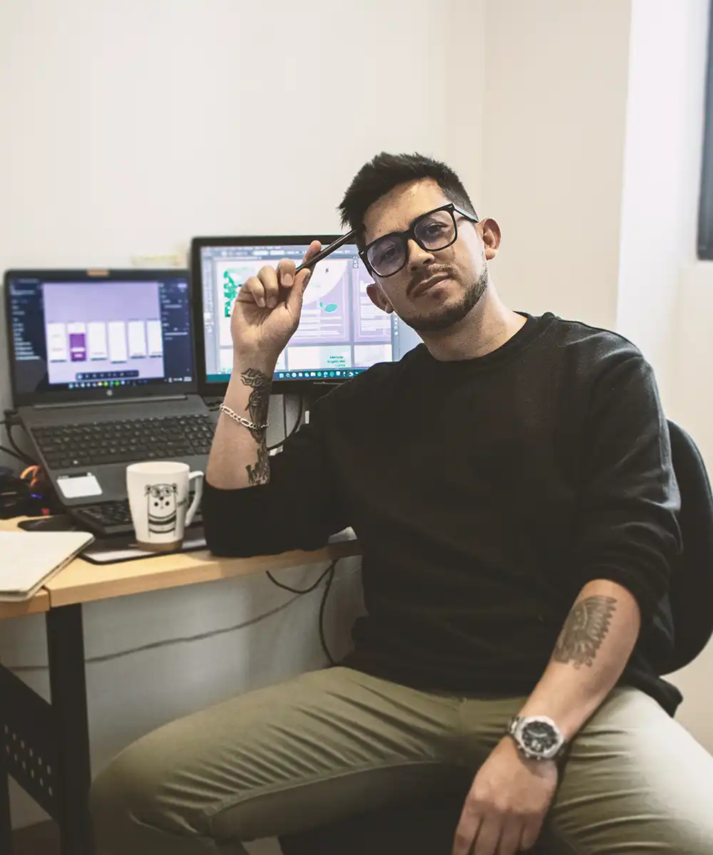 Un hombre joven con barba, bigote y gafas, sentado en su escritorio. Viste una sudadera negra y pantalones de color caqui, y tiene varios tatuajes en los brazos. Sostiene un lápiz con la mano derecha y mira directamente a la cámara. Detrás de él se ven dos monitores de computadora con gráficos y diseños. Representa a una persona en su espacio de trabajo creativo.
